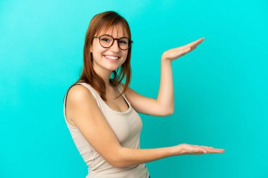 Redhead girl isolated on blue background holding copyspace to insert an ad