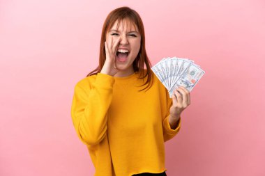 Redhead girl taking a lot of money isolated on pink background shouting with mouth wide open