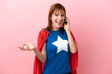 Super Hero redhead girl isolated on pink background keeping a conversation with the mobile phone with someone