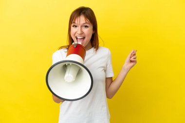 Redhead girl isolated on yellow background shouting through a megaphone and pointing side