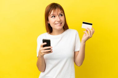 Redhead girl isolated on yellow background buying with the mobile with a credit card while thinking