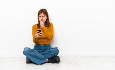 Redhead girl sitting on the floor isolated on white background thinking and sending a message