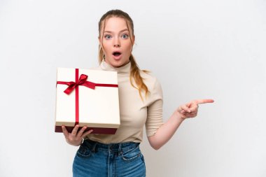 Young caucasian woman holding a gift isolated on white background surprised and pointing side