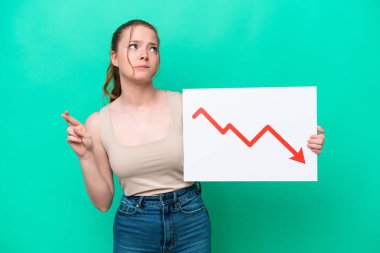 Young caucasian woman isolated on green background holding a sign with a decreasing statistics arrow symbol with fingers crossing