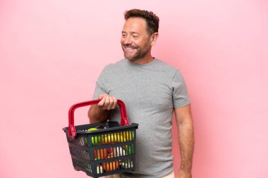 Orta yaşlı beyaz bir adam pembe arka planda izole edilmiş bir alışveriş sepeti tutuyor. Yan tarafa bakıyor ve gülümsüyor.