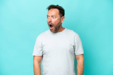 Mavi arka planda izole edilmiş orta yaşlı beyaz bir adam yan tarafa bakarken sürpriz jest yapıyor.