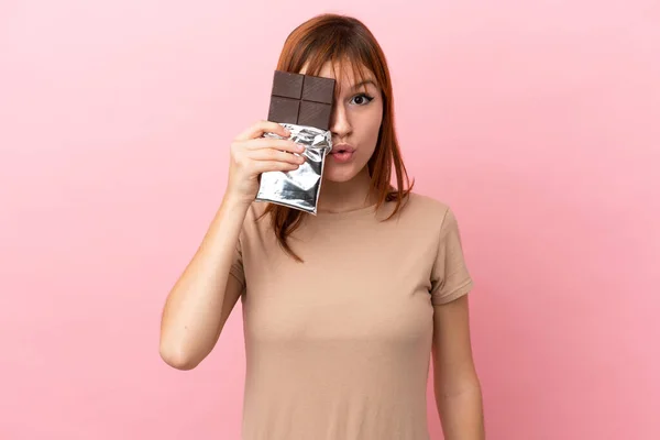 Redhead Girl Isolated Pink Background Taking Chocolate Tablet Surprised — Stockfoto