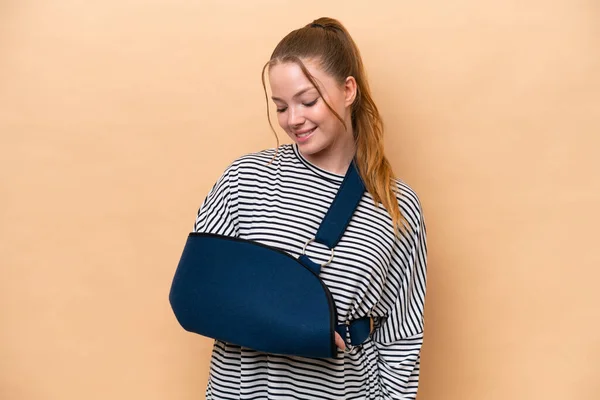 Young caucasian girl with broken arm and wearing a sling isolated on beige background with happy expression