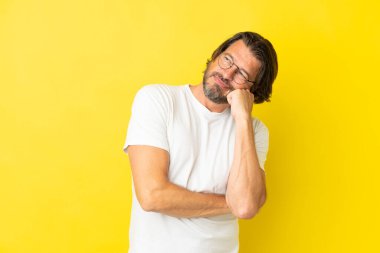 Yaşlı Hollandalı adam, yorgun ve sıkılmış ifadesiyle sarı arka planda izole edilmiş.