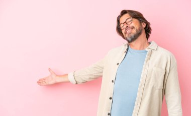 Kıdemli Hollandalı, pembe arka planda izole edilmiş. Davet etmek için ellerini yana uzatıyor.