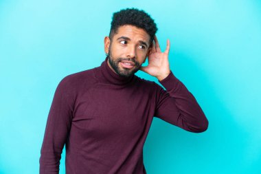 Genç Brezilyalı bir adam mavi arka planda izole edilmiş bir şekilde elini kulağına koyarak bir şeyler dinliyor.
