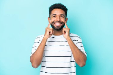 Genç Brezilyalı adam mavi arka planda izole edilmiş mutlu ve hoş bir ifadeyle gülümsüyor.