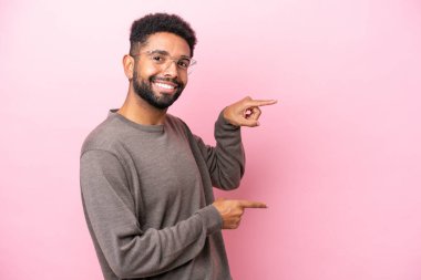 Pembe arka planda izole edilmiş genç Brezilyalı bir adam parmak göstererek bir ürün sunuyor.