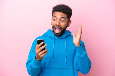 Pembe arka planda izole edilmiş genç Brezilyalı adam kameraya bakarken şaşırmış bir ifadeyle cep telefonuna bakıyor.