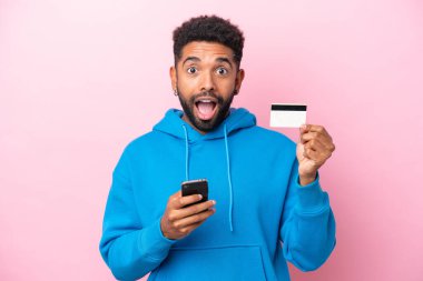 Genç Brezilyalı bir adam pembe arka planda izole edilmiş cep telefonuyla alışveriş yapıyor ve elinde şaşırmış bir ifadeyle bir kredi kartı tutuyor.