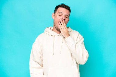 Beyaz tenli yakışıklı adam mavi arka planda izole edilmiş esniyor ve ağzı açık bir şekilde elleriyle ağzını kapatıyor.