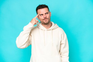Genç, beyaz, yakışıklı bir adam mavi arka planda izole edilmiş. Delilik jesti yapıyor. Parmağını kafaya koyuyor.
