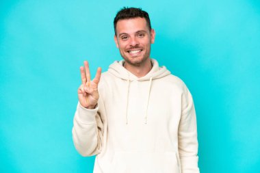 Beyaz tenli yakışıklı adam mavi arka planda izole edilmiş mutlu ve parmaklarıyla üçe kadar sayıyor.
