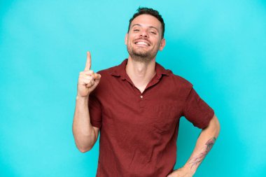 Beyaz tenli yakışıklı adam mavi arka planda izole edilmiş ve en iyinin işareti olarak parmağını kaldırıyor.