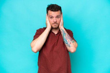 Kafkasyalı yakışıklı adam mavi arka planda izole edilmiş hayal kırıklığına uğramış ve kulaklarını kapatmış.