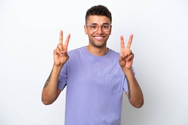 Beyaz arka planda izole edilmiş beyaz Brezilyalı genç adam iki eliyle zafer işareti gösteriyor.
