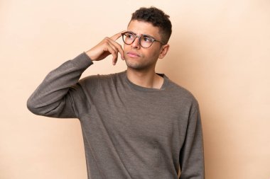 Genç Brezilyalı adam bej arka planda izole edilmiş. Delilik jesti yapıyor. Parmağını kafaya koyuyor.