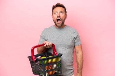 Elinde alışveriş sepeti olan orta yaşlı beyaz bir adam pembe arka planda izole edilmiş yukarı bakıyor ve şaşırmış bir ifadeyle