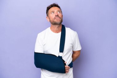 Middle age caucasian man with broken arm and wearing a sling isolated on purple background and looking up