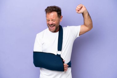 Kırık kollu orta yaşlı beyaz bir adam ve mor arka planda izole edilmiş askısı var. Güçlü bir hareket yapıyor.