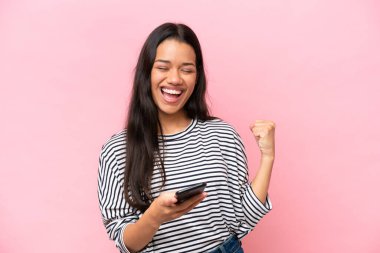 Pembe arka planda izole edilmiş genç Kolombiyalı kadın telefon zafer pozisyonunda.