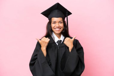 Kolombiyalı genç bir üniversite mezunu. Pembe arka planda izole edilmiş. Baş parmakları havada ve gülümsüyor.