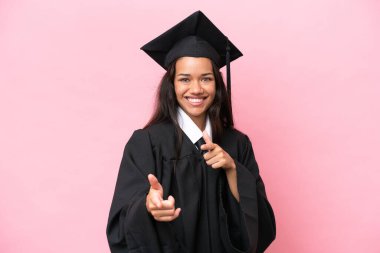 Kolombiyalı genç bir üniversite mezunu. Pembe arka planda izole edilmiş. Ön tarafı işaret ediyor ve gülümsüyor.