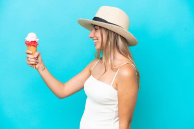 Mavi arka planda izole edilmiş kornet dondurmalı mayolu beyaz kadın mutlu bir ifadeyle.