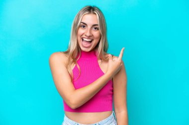 Mavi arka planda izole edilmiş genç beyaz kadın şaşırmış ve işaret ediyor.