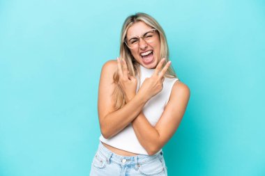 Mavi arka planda izole edilmiş genç beyaz kadın gülümsüyor ve zafer işareti gösteriyor.