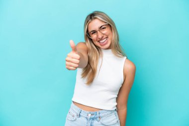 Genç beyaz kadın mavi arka planda izole edildi. Başparmakları havada çünkü iyi bir şey oldu.