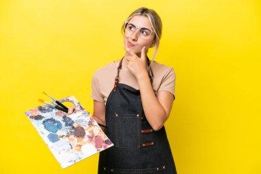 Young artist caucasian woman holding a palette isolated  on yellow background having doubts