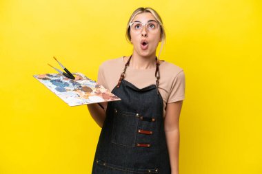 Sarı arka planda izole edilmiş palet tutan beyaz bir kadın yukarı bakıyor ve şaşırmış bir ifadeyle
