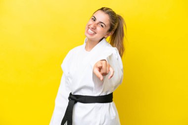 Sarı arka planda karate yapan genç beyaz kadın mutlu bir ifadeyle öne bakıyor.