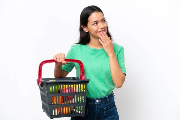 白い背景に隔離された食べ物でいっぱいの買い物かごを持っている若いコロンビア人女性は笑顔で見上げます — ストック写真