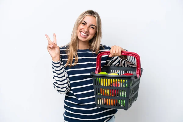 年轻的高加索女人拿着一个装满食物的购物篮 被白色背景隔离 面带微笑地展示胜利的标志 — 图库照片