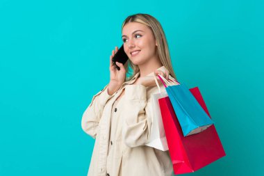 Kafkasyalı genç bir kadın mavi arka planda izole edilmiş. Elinde alışveriş poşetleri var ve cep telefonuyla arkadaşını arıyor.