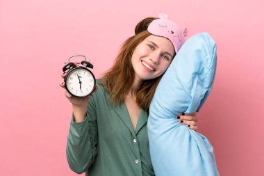 Pijamalı genç İngiliz kadın pembe arka planda pijamalarıyla izole edilmiş ve elinde mutlu bir ifadeyle saat ve yastık tutuyordu.