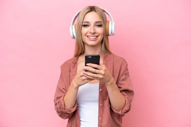 Güzel sarışın kadın pembe arka planda izole edilmiş. Cep telefonuyla müzik dinliyor ve ön tarafa bakıyor.