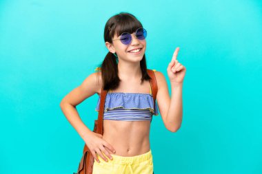 Küçük beyaz bir çocuk kumsala gidiyor. Mavi arka planda izole edilmiş. En iyinin işareti olarak parmağını kaldırıyor.