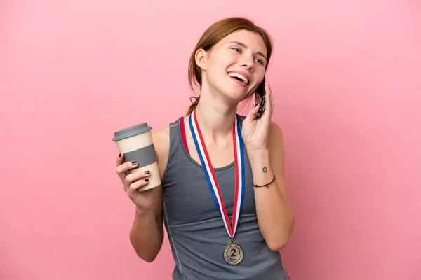 Young English woman with medals isolated on pink background holding coffee to take away and a mobile