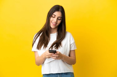 Sarı arka planda izole edilmiş genç beyaz kadın cep telefonuyla bir mesaj gönderiyor.