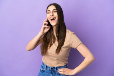 Mor arka planda izole edilmiş cep telefonu kullanan beyaz bir kadın. Kolları kalçasında ve gülümsüyor.
