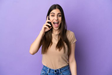 Mor arka planda izole edilmiş cep telefonu kullanan beyaz bir kadın. Şaşırtıcı bir yüz ifadesi var.