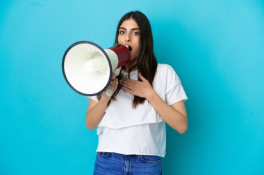 Young caucasian woman isolated on blue background shouting through a megaphone with surprised expression